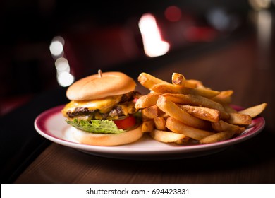 Iconic Diner Double Cheeseburger Of Stacked Juicy Beef Patty And Melted Cheese On Lettuce And Tomato Between Soft Potato Buns With Thick Steak Cut Golden French Fries And Dark Booth Blurry Background