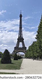 Iconic Eiffel Tower stands majestically at Champ de Mars, Paris, framed by green trees and a walking path under bright blue sky. Symbolizes travel, romance, and European architecture on a sunny day.