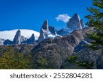 The iconic Fitz Roy and Aguja Poincenot peaks rise majestically against a vivid blue sky, surrounded by rugged terrain and greenery in El Chaltén, Patagonia, Argentina.