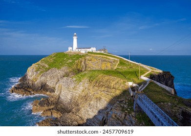 An iconic view of the South Stack Lighthouse at sunrise perched on a rugged island cliff off the coast of Anglesey, North Wales, UK. The image features the white lighthouse tower, the surrounding grassy rock formations, and the metal footbridge, set against a calm blue sea and a clear sky, perfect for travel, maritime, and landscape photography.