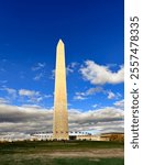 Iconic view of the Washington Monument under a vivid blue sky with dramatic clouds. Includes rows of American flags encircling the base, capturing the grandeur of this historic landmark.