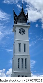 An iconic white clock tower featuring traditional Minangkabau architecture stands majestically beneath a vibrant blue tropical sky and dramatic white clouds. Captured from a low-angle perspective, this stunning cultural landmark showcases the rich heritage, unique design, and architectural beauty of West Sumatra, Indonesia, making it a captivating destination for travelers, culture enthusiasts, and architecture lovers alike.