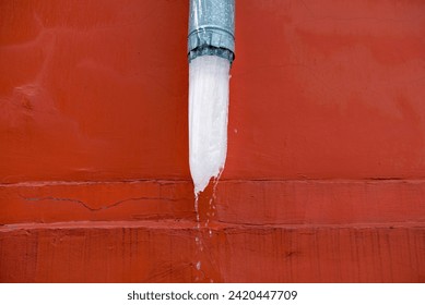 Icy drain pipes on red facade of the building. Ice block thaw in rain gutter. Gutter downspout with melting ice. Icicles melt in gutter. Winter ice melt in drain system. Drain pipe with ice