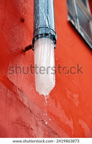 Icy drain pipes on red facade of the building. Ice block thaw in rain gutter. Downspout with melting ice. Icicles melt in gutter. Winter ice melt in drain system. Drain pipe with ice, Selective focus