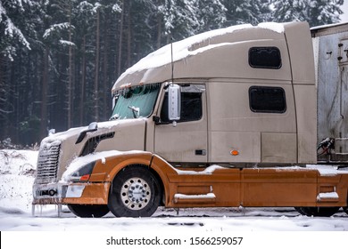 Icy frozen dirty brown big rig bonnet powerful industrial grade semi truck for long haulage with dry van semi trailer standing on the winter parking lot with ice, snow and trees