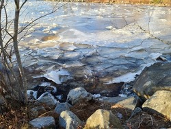 An Icy Shoreline Alongside Of A Pond. The Water Is Completely Frozen Over And The Ice Is Chipping Around The Jagged Edged Boulders.