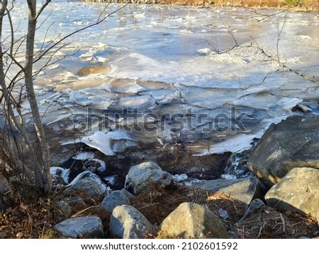 An icy shoreline alongside of a pond. The water is completely frozen over and the ice is chipping around the jagged edged boulders.