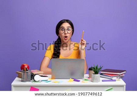 Idea Concept. Excited Young Indian Woman In Eyeglasses Sitting At The Desk Using Laptop And Raising Pencil Up Isolated Over Purple Background In Studio. Amazed Shocked Lady Having Insight, Aha Moment.