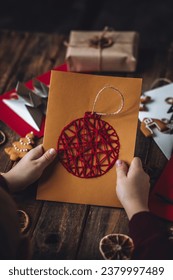 Idea for easy Christmas do it yourself crafts for kids to make. Child's hands with handmade postcards with fir tree and toy from recycled cardboard paper, thread, buttons. Wooden background, flat lay
