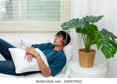 The idea of ​​relaxing at home. A handsome Asian man wears headphones while lying on the sofa listening to music in his apartment. Meditation or podcast with eyes closed. relax in the bedroom - Powered by Shutterstock