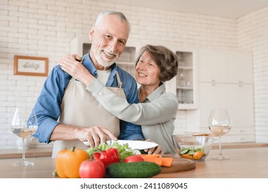 Ideal happy senior couple. Old elderly wife hugging embracing cuddling with husband, helping him prepare dinner, cooking vegetable salad together - Powered by Shutterstock