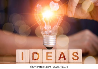 Ideas creativity concept, hand touching light bulb and ideas wording on wood cube square on table. 