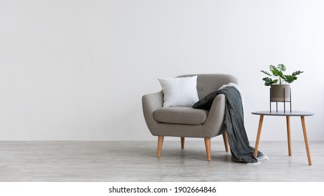 Ideas for modern minimal home interior design. Gray vintage armchair with white pillow and blanket, table with green plant in pot on floor, on light wall background, panorama, copy space, nobody