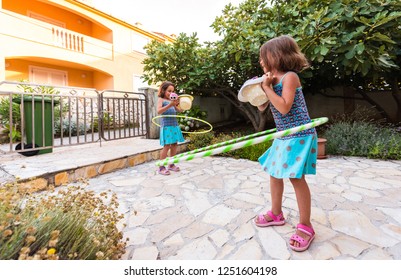 Identical twin sisters are playing on vacation with hula hoop. Sisters wearing blue dresses and hats are enjoying vacation hooping and laughing on stone paved floor front yard of vacation home. - Powered by Shutterstock