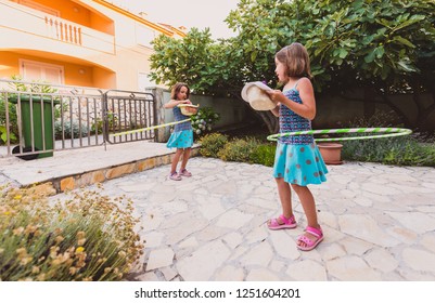 Identical twin sisters are playing on vacation with hula hoop. Sisters wearing blue dresses and hats are enjoying vacation hooping and laughing on stone paved floor front yard of vacation home. - Powered by Shutterstock