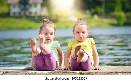 Identical Twins Girls Are Exercising On A Lake Shore. There Is Sun In The Background. Children  Physical Exercise And Healthy Lifestyle. Abdominal Training Or Workout In Nature