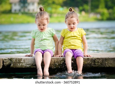 Identical Twins Girls Are Exercising On Lake Shore, Sprinkling Water. Children Sitting On Lake Side, Playing With Water. Healthy And Active Children Lifestyle.