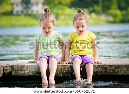 Identical twins girls are exercising on lake shore, sprinkling water. Children sitting on lake side, playing with water. Healthy and active children lifestyle.