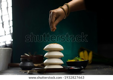 Idly or Idli, south indian breakfast item which is placed one over other beautifully and a woman hand garnishing idli with chilli flakes which is in a black plate  on dark textured background.