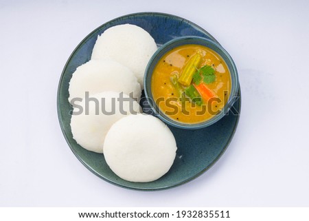 Idly or Idli, south indian main breakfast item which is beautifully arranged in aqua colour plates with a small bowl of sambar placed on white background.