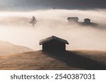 Idyllic alpine meadow during a moody, atmospheric and misty sunrise in Alpe di Siusi with rustic huts and landscape views of the Dolomite mountains in South Tyrol, Italy.