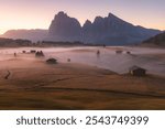 Idyllic alpine meadow during a moody, atmospheric and misty sunrise in Alpe di Siusi with rustic huts and landscape views of the Dolomite mountains in South Tyrol, Italy.