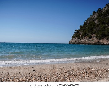 Idyllic beach scene with a pebbled shore, turquoise waves, and a rugged cliffside covered in trees under a clear blue sky, showcasing natural coastal beauty. - Powered by Shutterstock