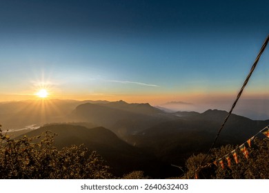 An idyllic landscape featuring the sun setting behind the majestic mountain range, visible through a cable suspension bridge