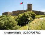 Idyllic low-angle view of castle Castelo de Aljezur with its tower and walls surrounded by green grass and bushes, featuring a waving Portuguese flag against the clear blue sky on a sunny spring day.