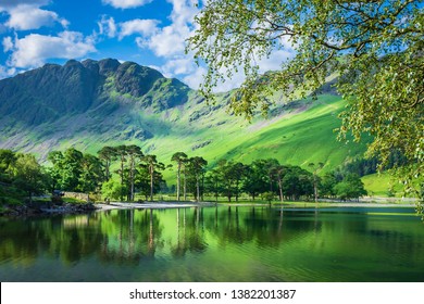 Idyllic scenery of English Lake District in spring.Trees growing on lakeshore and green hill reflecting in lake water.Tree branches in foreground and mountain peak in background.Majestic landscape.