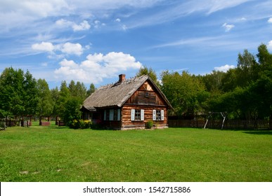 An Idyllic View Of An Old Rural Rustic House With A Slanted Thatch Roof And A Chimney Standing In The Middle Of A Freshly Mown Field Or Lawn Next To An Orchard And A Dense Moor On A Cloudy Summer Day