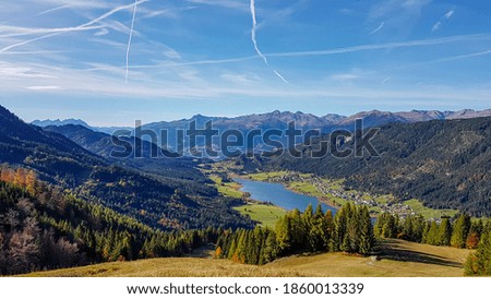 An idyllic view from the side of the mountain on a distant Weissensee lake in Austria. The grass is golden, trees also change colors to yellow and orange. Autumn vibes. High mountains around. Calmness