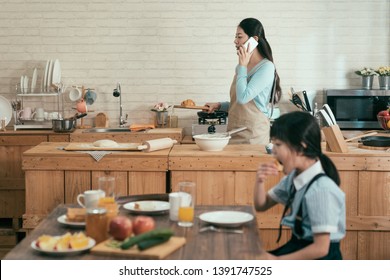 Ignore Family Gadget Addiction. Busy Asian Wife Mother Holding Wooden Plate With Croissant Walking In Modern Kitchen Talking With Husband On Cellphone In Morning. Little Girl Eating Having Breakfast