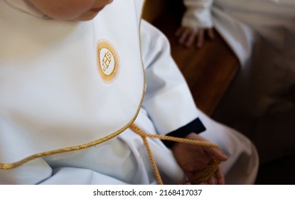 IHS Christian symbol on a communion child. IHS Christogram embroidered with gold thread on a white alba. First Holy Communion dress.