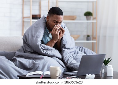 Ill African Man Having Rhinitis Sneezing And Blowing Runny Nose In Paper Tissue Sitting On Sofa At Home, Working Distantly On Laptop. Sinusitis Illness, Cold And Influenza Symptoms.