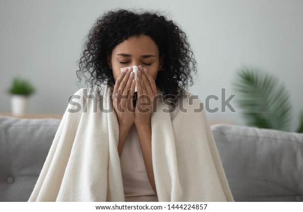 Ill african young woman covered with blanket\
blowing running nose got fever caught cold sneezing in tissue sit\
on sofa, sick allergic black girl having allergy symptoms coughing\
at home, flu concept