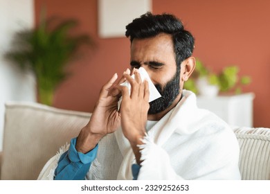 Ill arabic guy sneezing and blowing runny nose in paper tissue sitting covered with blanket on sofa at home. Portrait of sick young man suffering from sinusitis illness, cold and influenza symptoms - Powered by Shutterstock