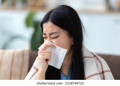 Ill Asian young woman sitting on the couch at home covered with blanket, blowing nose, feeling unwell, has a cold or flu - Shutterstock ID 2118744713
