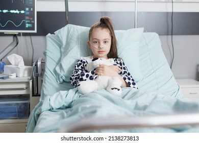 Ill little girl holding plush teddybear and wearing oxygen tube while resting alone in hospital bed. Unwell child in healthcare pediatric clinic patients recovery ward room while looking at camera.