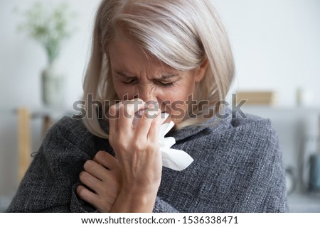 Ill mature woman covered with blanket blowing running nose sneeze in tissue suffer from allergy flu, allergic old lady hold handkerchief got hay fever rhinitis symptom cough at home, allergy concept
