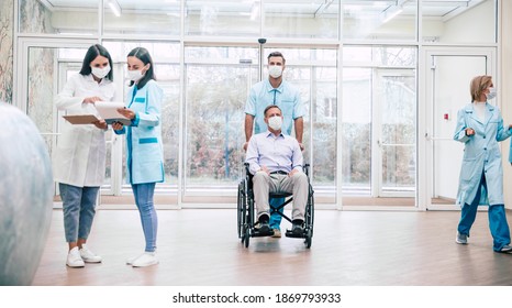 Ein älterer Mann mit Schutzmaske auf dem Gesicht in einem Rollstuhl und ein selbstbewusster Arzt in der medizinischen Maske während des Transports im Krankenhaus.