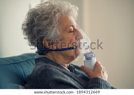 Ill senior woman making inhalation at home. Close up of an elderly woman holding mask nebulizer inhaling fumes medication into lungs. Self treatment of the respiratory tract using inhalation nebulizer