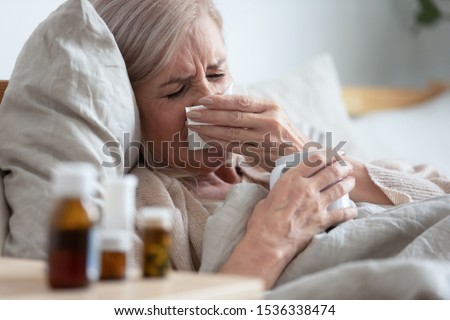 Ill sick middle aged woman sneezing blowing running nose holding tissue sit on bed, upset old mature lady caught cold got flu influenza grippe symptoms drink hot tea taking medications at home alone