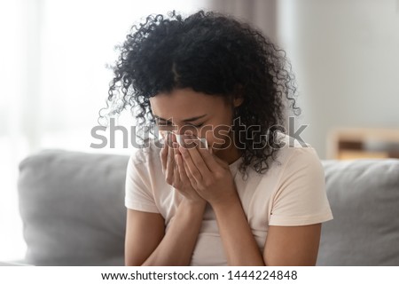 Ill upset african woman blowing running nose got flu caught cold sneezing in tissue sit on sofa, sick allergic black girl having allergy symptoms coughing holding napkin at home, hay fever concept