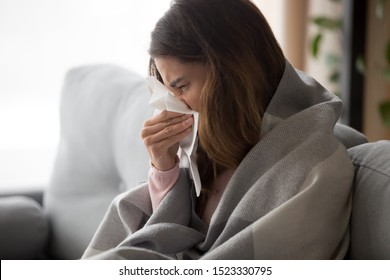 Ill upset young woman sitting on sofa covered with blanket freezing blowing running nose got fever caught cold sneezing in tissue, sick girl having influenza symptoms coughing at home, flu concept - Shutterstock ID 1523330795