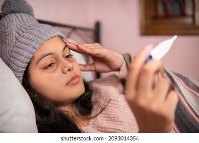 Ill woman lying on the bed having high fever and measuring her temperature with a thermometer and touching her forehead. She is wearing warm clothes.