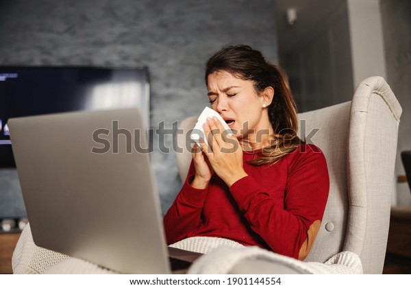 Ill woman sitting in chair at home\
during lockdown with laptop in her lap and\
sneezing