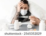 Ill young man lying under blanket on bed and showing thermometer with temperature result to camera. A lot of pills on table. Covid-19 coronavirus disease, seasonal flu and cold, illness fatigue