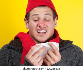 Ill young man with red nose, scarf and cap sneezing into handkerchief
