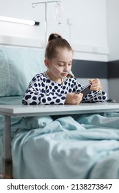 Ill Young Patient Playing On Smartphone While Resting On Bed In Pediatric Hospital Room. Sick Joyful Young Girl Enjoying Mobile Phone Entertainment While Sitting In Clinic Recovery Ward.